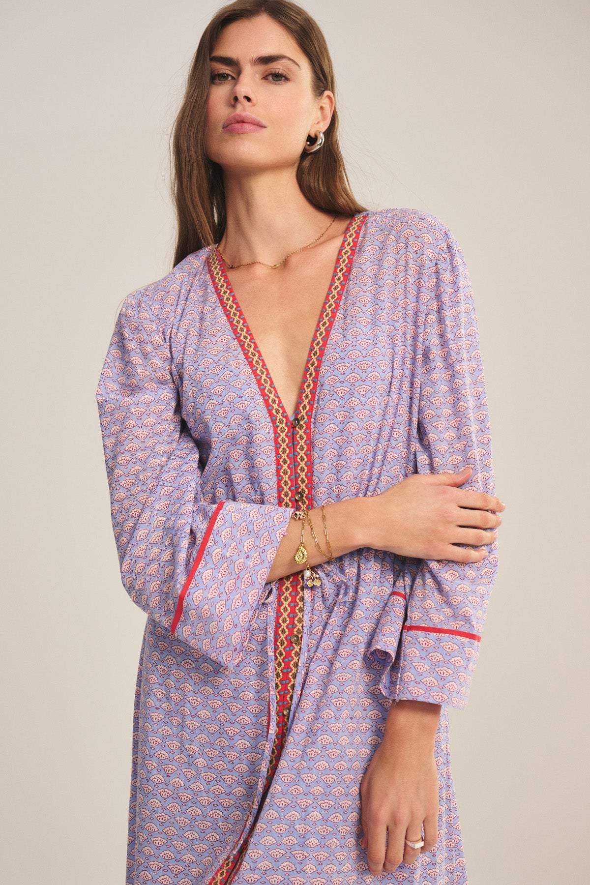 A woman stands against a plain background wearing the WRENLEY DRESS by Velvet by Graham & Spencer, a patterned, long-sleeved midi dress featuring a deep V-neckline, red trim, paisley print, and delicate embroidered detail.