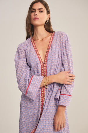 A woman stands against a plain background wearing the WRENLEY DRESS by Velvet by Graham & Spencer, a patterned, long-sleeved midi dress featuring a deep V-neckline, red trim, paisley print, and delicate embroidered detail.