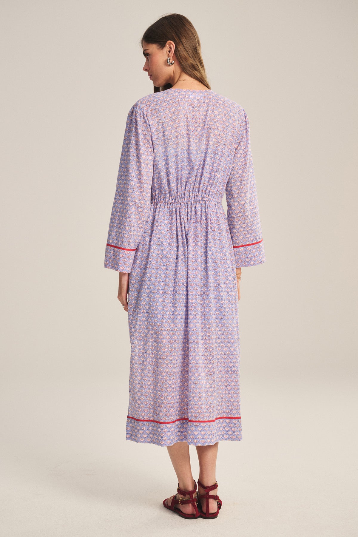 A woman faces away, wearing the WRENLEY DRESS by Velvet by Graham & Spencer—a long-sleeved cotton voile midi with red trim and embroidered details—paired with red sandals against a neutral backdrop.