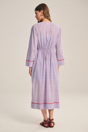 A woman faces away, wearing the WRENLEY DRESS by Velvet by Graham & Spencer—a long-sleeved cotton voile midi with red trim and embroidered details—paired with red sandals against a neutral backdrop.