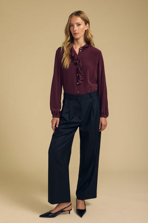 A woman stands against a beige background wearing the PAIGE SILK BLOUSE by Velvet by Graham & Spencer—a burgundy washed silk top with a ruffle neckline—paired with black wide-leg trousers and black slingback shoes.