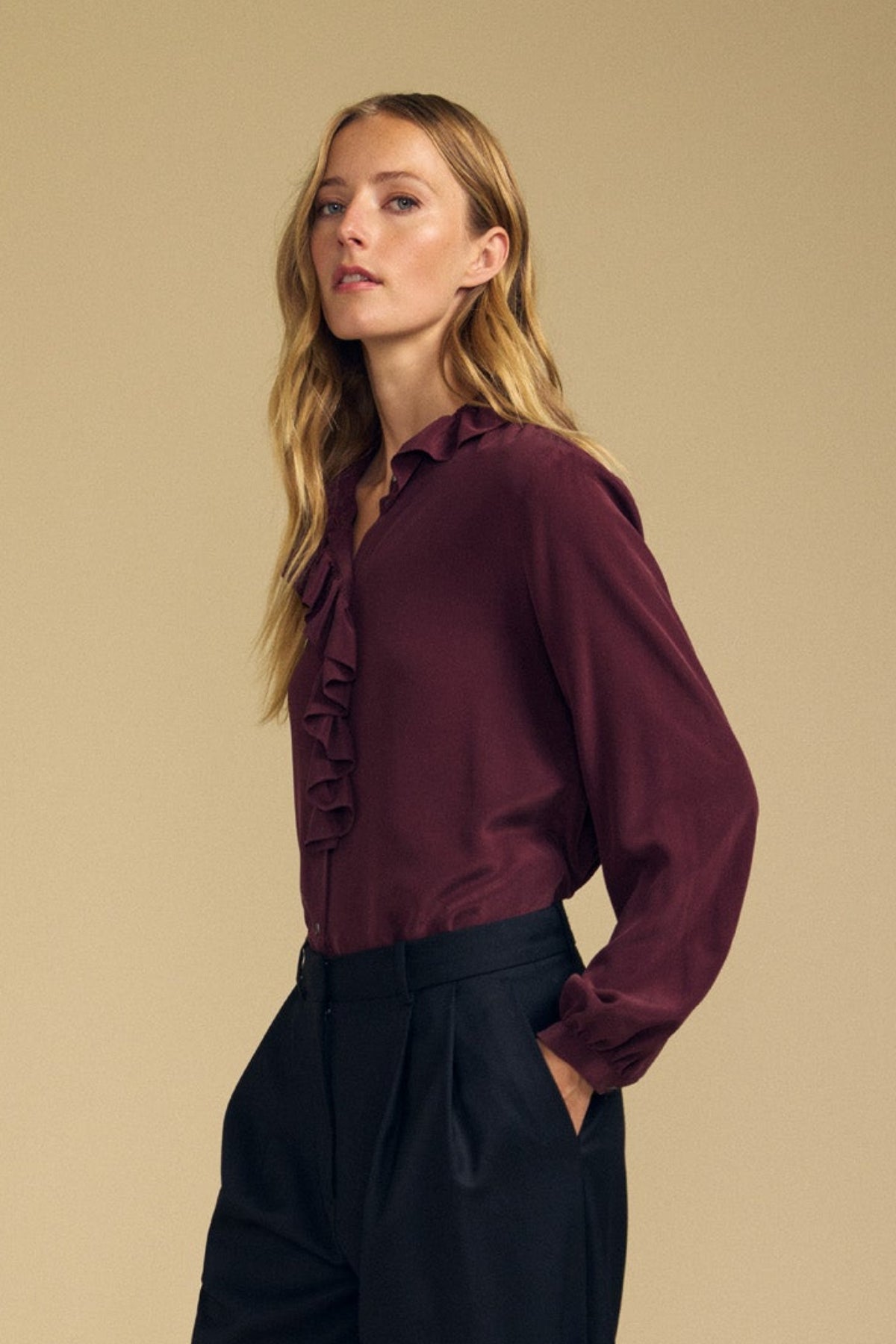 A woman with long blonde hair wears the PAIGE SILK BLOUSE by Velvet by Graham & Spencer—featuring a washed silk burgundy fabric and ruffle neckline—paired with black pants, standing against a plain beige background.-42063833956545