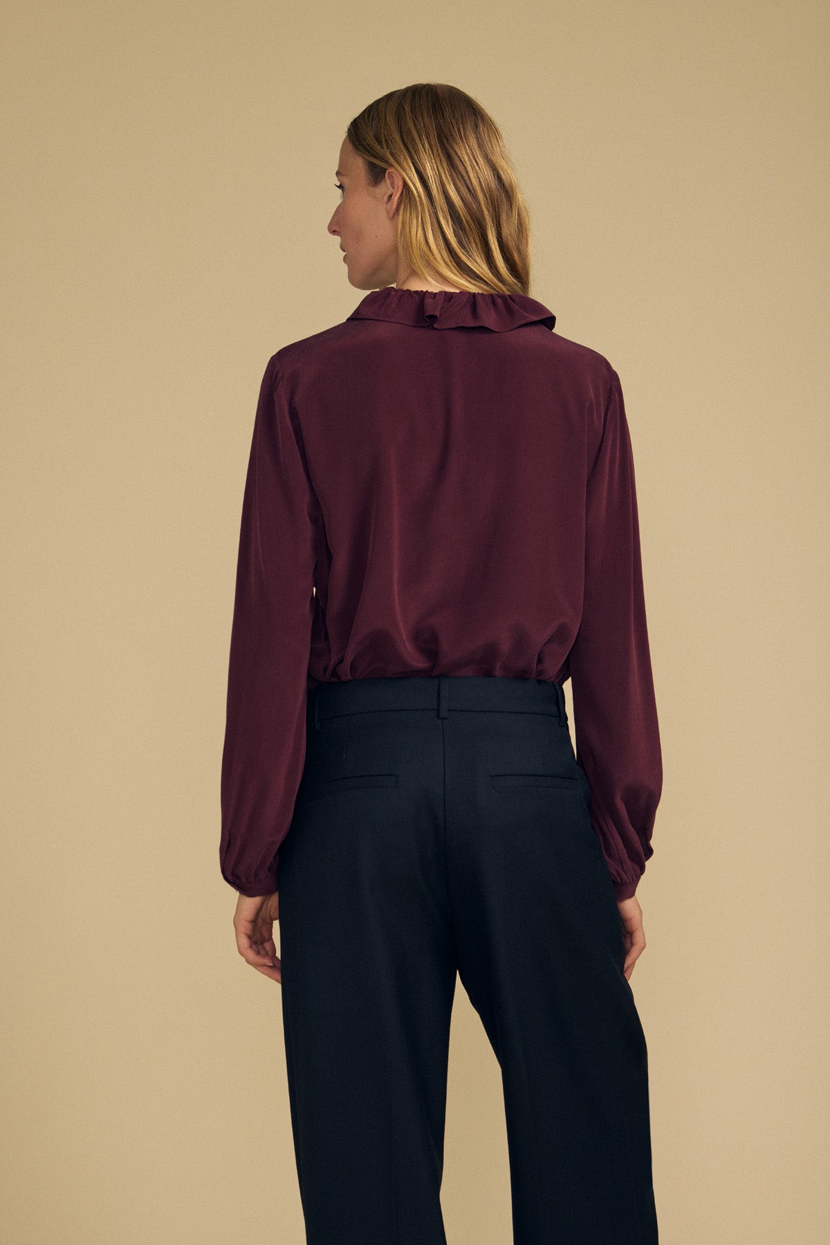 A person stands with their back to the camera, wearing the PAIGE SILK BLOUSE by Velvet by Graham & Spencer—a burgundy washed silk top with a ruffle neckline—paired with black trousers against a plain beige background.-42063834087617