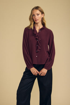 A woman stands against a beige background wearing the Velvet by Graham & Spencer PAIGE SILK BLOUSE—a long-sleeve, burgundy washed silk top with a ruffle neckline—paired with black wide-leg pants.