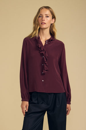 A woman stands against a plain beige background, wearing the PAIGE SILK BLOUSE by Velvet by Graham & Spencer—a long-sleeve maroon washed silk blouse featuring a ruffle neckline and a matte finish—paired with black pants.