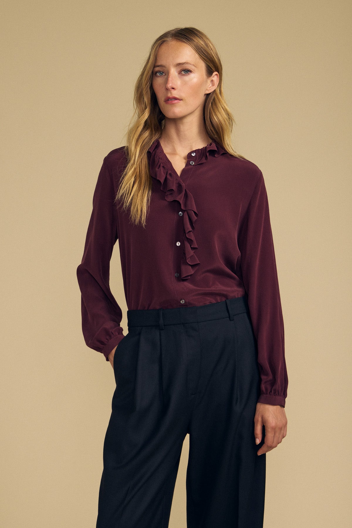 A woman stands against a beige background wearing the PAIGE SILK BLOUSE by Velvet by Graham & Spencer—a burgundy washed silk blouse featuring a ruffle neckline—paired with black trousers, with one hand in her pocket.