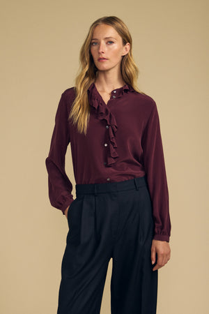 A woman stands against a beige background wearing the PAIGE SILK BLOUSE by Velvet by Graham & Spencer—a burgundy washed silk blouse featuring a ruffle neckline—paired with black trousers, with one hand in her pocket.