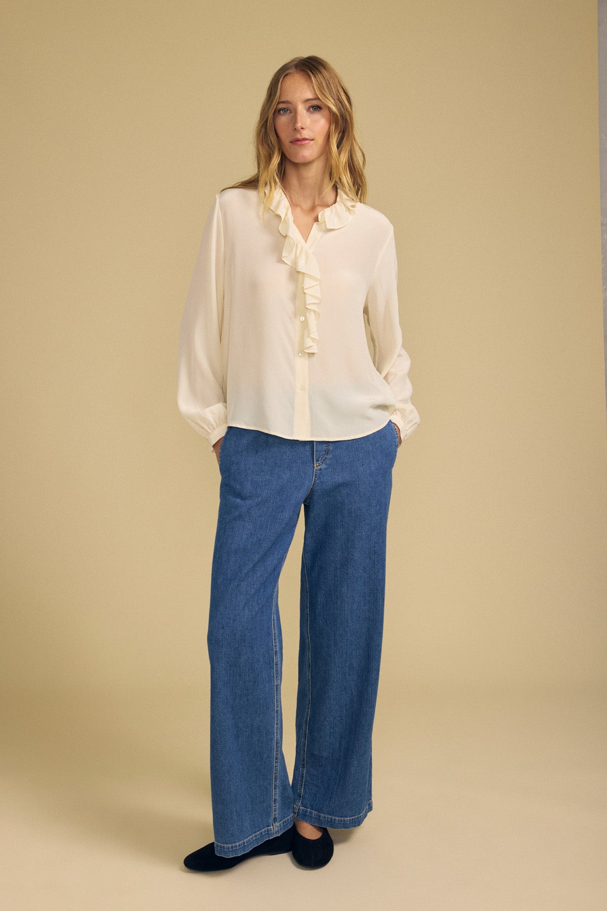 A woman stands against a beige background wearing the PAIGE SILK BLOUSE by Velvet by Graham & Spencer, featuring a ruffle neckline, paired with wide-leg blue jeans and black shoes, her hands in her pockets.-42063834153153