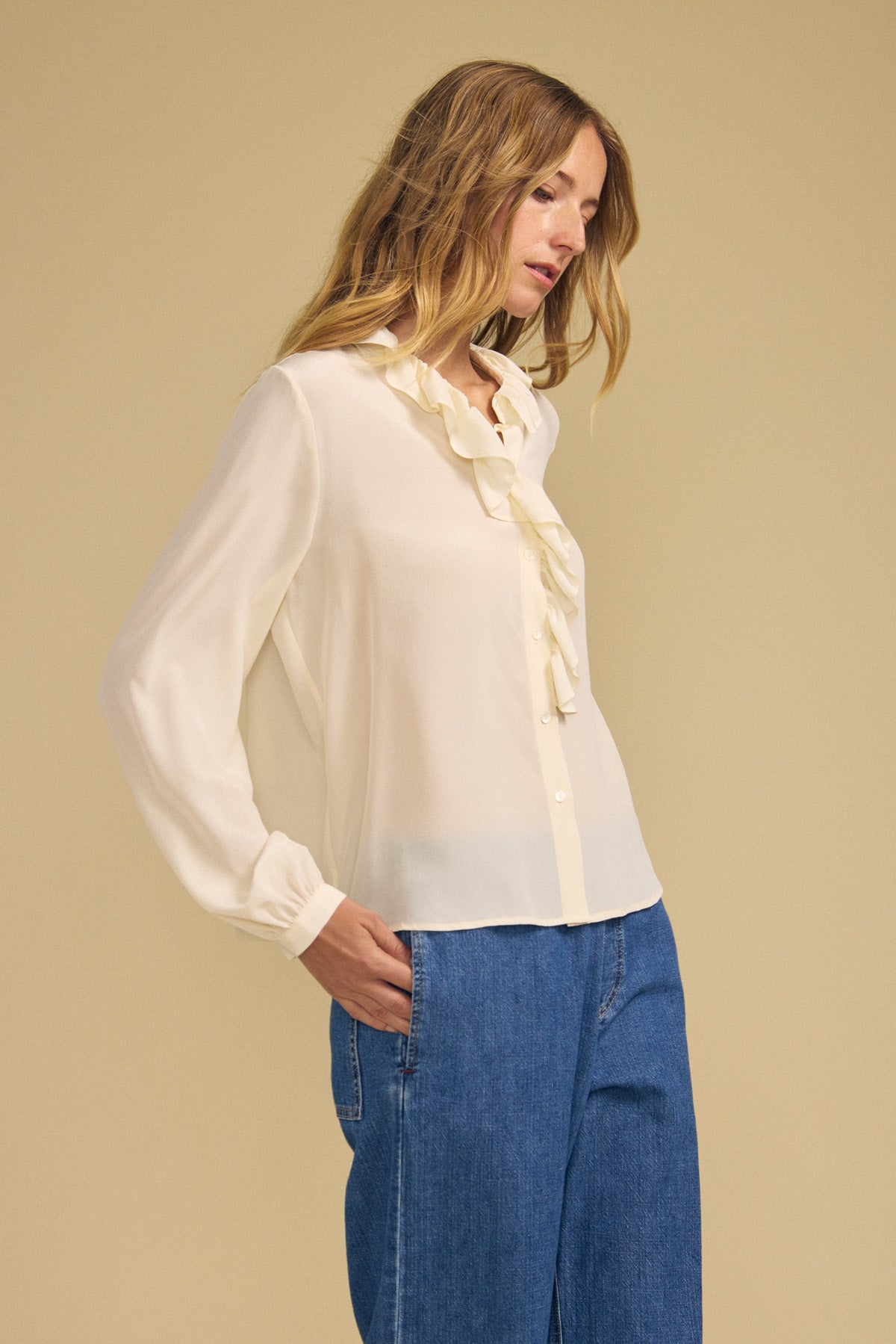 A woman wearing the PAIGE SILK BLOUSE by Velvet by Graham & Spencer—a cream washed silk blouse with a ruffle neckline—paired with blue jeans, standing against a beige background.