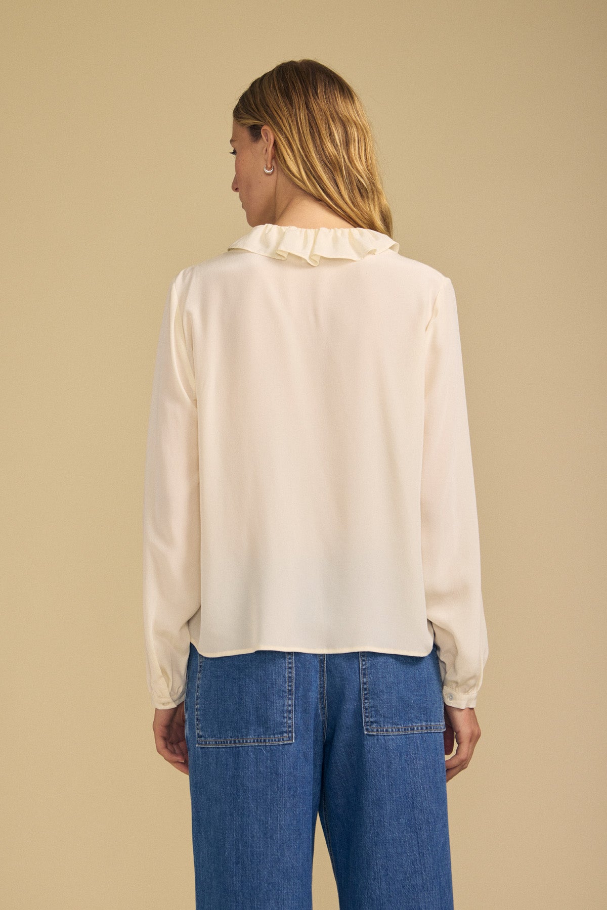 A woman with long hair faces away, wearing the PAIGE SILK BLOUSE by Velvet by Graham & Spencer—a cream-colored washed silk top with a ruffle neckline—paired with blue jeans, standing against a plain beige background.