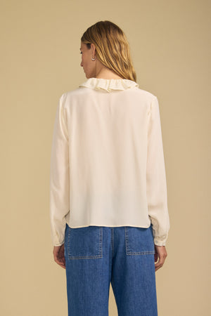 A woman with long hair faces away, wearing the PAIGE SILK BLOUSE by Velvet by Graham & Spencer—a cream-colored washed silk top with a ruffle neckline—paired with blue jeans, standing against a plain beige background.