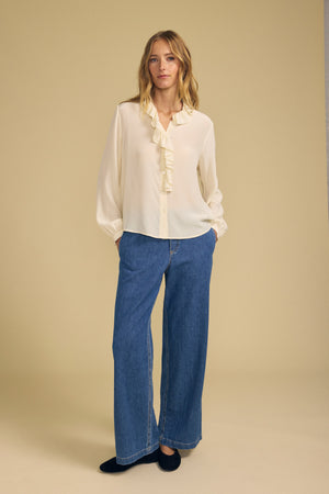 A woman stands against a beige background wearing the PAIGE SILK BLOUSE by Velvet by Graham & Spencer, featuring a ruffle neckline, paired with wide-leg blue jeans and black shoes, her hands in her pockets.