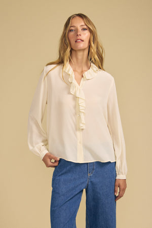 A person wearing the PAIGE SILK BLOUSE by Velvet by Graham & Spencer, a cream washed silk top with a ruffle neckline and a subtle matte finish, pairs it with blue jeans while standing against a plain beige background, facing the camera with a neutral expression. The elegant sheen of the blouse enhances the overall look.
