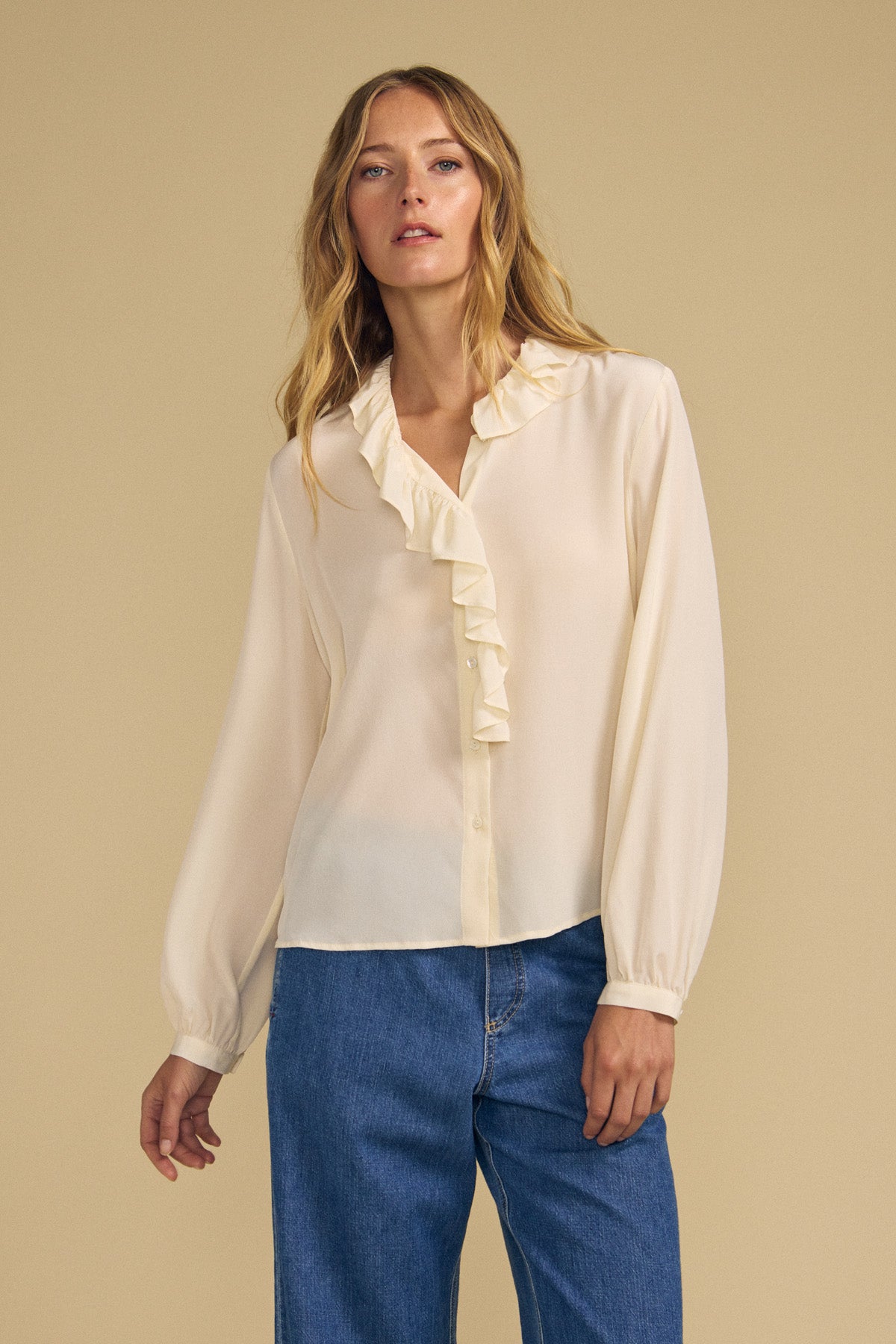 A woman wears the PAIGE SILK BLOUSE by Velvet by Graham & Spencer—a cream-colored washed silk top featuring a ruffled neckline and long sleeves—paired with blue jeans, standing against a plain beige background.-42063834349761