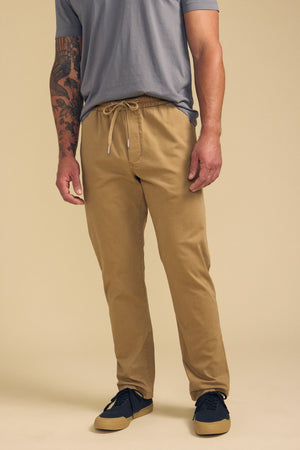 A man wearing a light grey t-shirt, Velvet by Graham & Spencer CONROY CHINO drawstring pants in tan, and black sneakers stands against a plain beige background.
