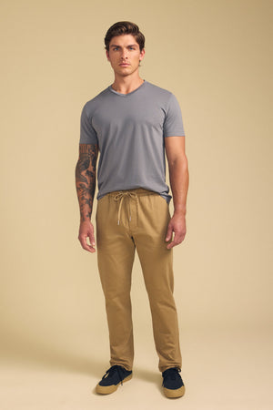 A man stands facing forward against a plain beige background, wearing Velvet by Graham & Spencer's CONROY CHINO drawstring pants, paired with a gray t-shirt and black shoes.