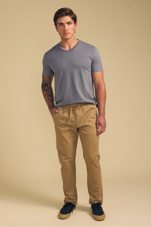 A man with a tattoo on his left arm, wearing Velvet by Graham & Spencer’s CONROY CHINO pants, a gray T-shirt, and black sneakers stands against a beige background with a neutral expression.