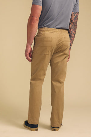 A man wearing tan CONROY CHINO pants by Velvet by Graham & Spencer and a light gray T-shirt stands facing away, displaying the back pockets; his right arm shows a tattoo.