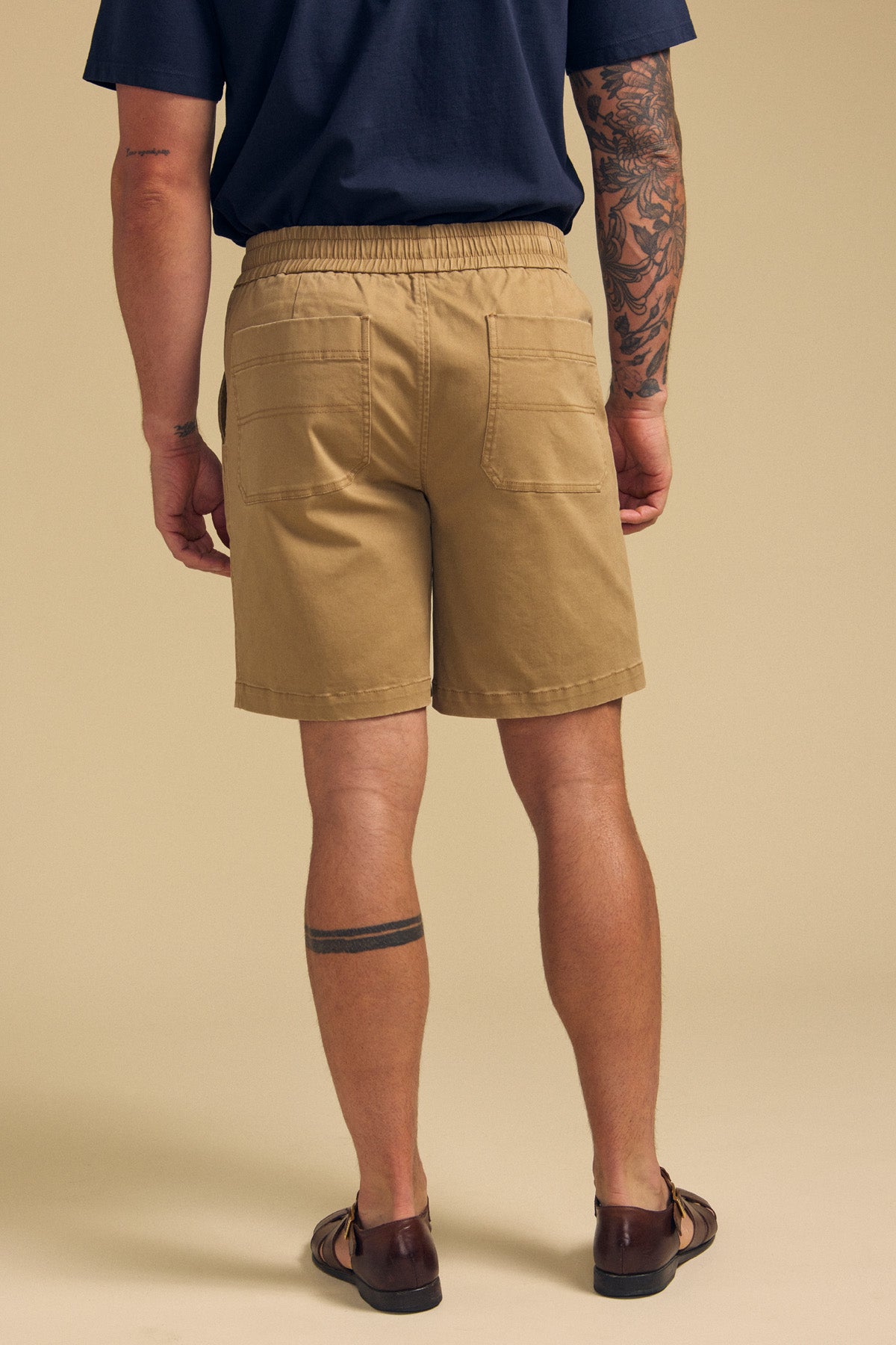 A person is shown from the back wearing tan GARTH SHORTs by Velvet by Graham & Spencer, a navy T-shirt, and brown sandals. Visible tattoos appear on their right arm and left calf against a neutral background.-41702010323137