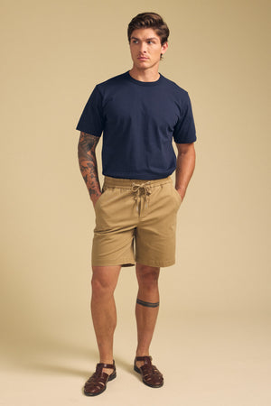 A man with tattoos on his left arm and right leg stands against a beige background, wearing navy tee, dark sandals, and tan GARTH SHORT from Velvet by Graham & Spencer.