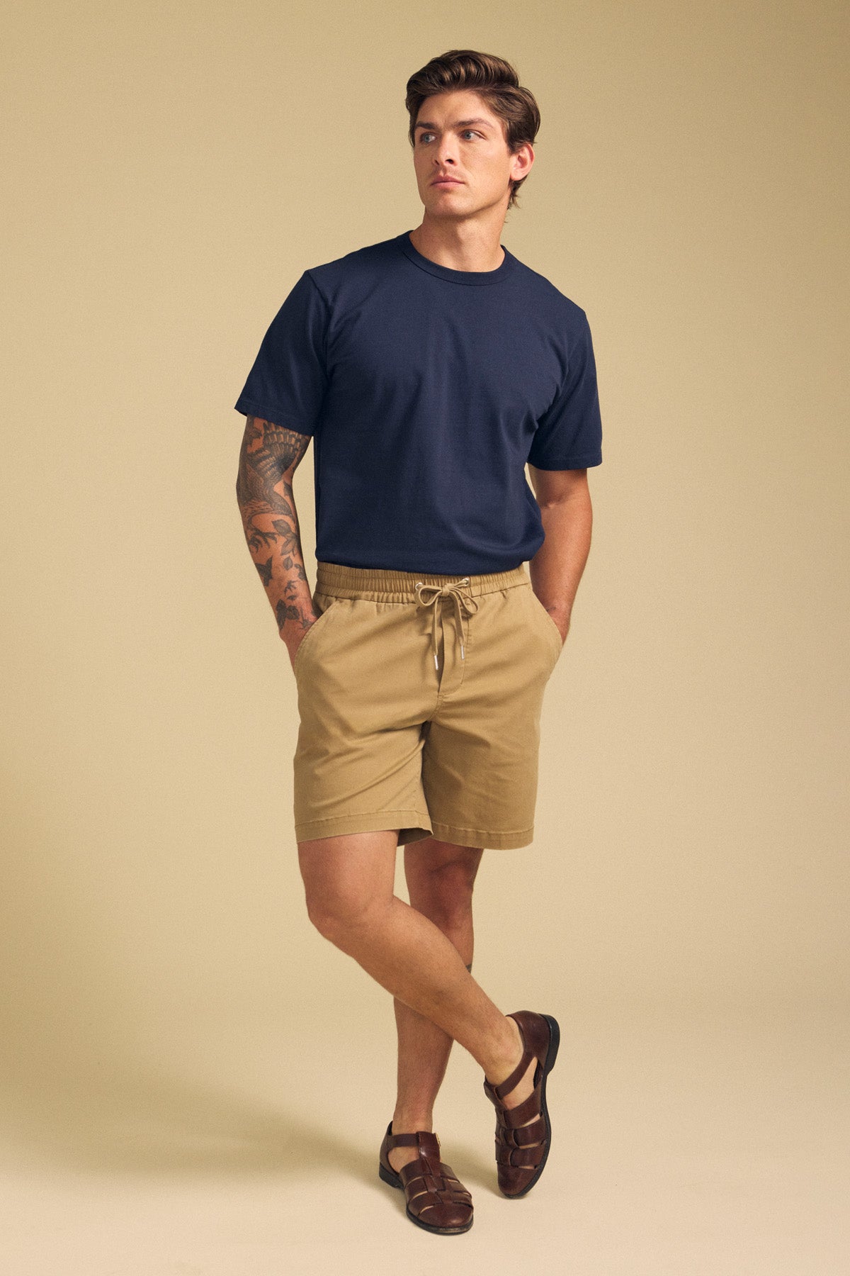 A man stands against a beige background wearing a navy t-shirt, Velvet by Graham & Spencer GARTH SHORT tan drawstring shorts, and brown sandals, with one hand in his pocket and visible arm tattoos.-41702010159297