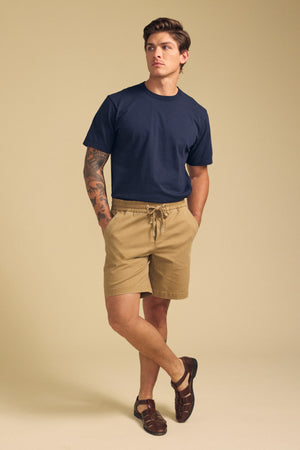 A man stands against a beige background wearing a navy t-shirt, Velvet by Graham & Spencer GARTH SHORT tan drawstring shorts, and brown sandals, with one hand in his pocket and visible arm tattoos.