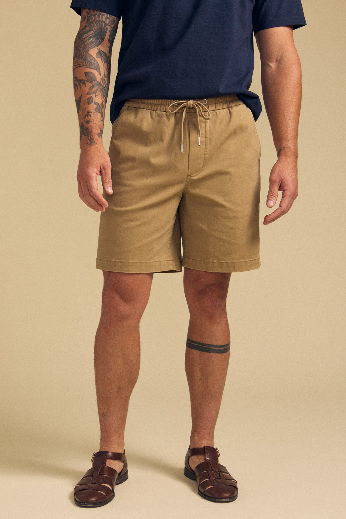 A person wears the GARTH SHORT by Velvet by Graham & Spencer in tan, paired with a navy t-shirt and brown sandals, showing tattoos on their left arm and right leg, standing against a plain beige background.