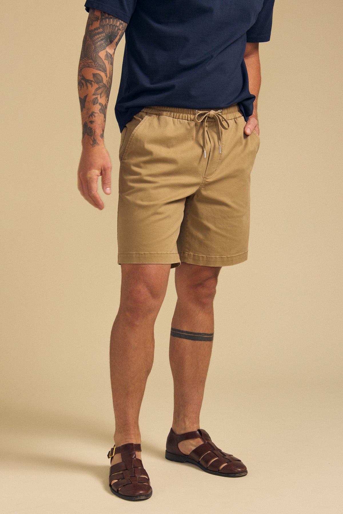 A person models Velvet by Graham & Spencer’s GARTH SHORT in tan, paired with a navy shirt and brown fisherman sandals, showing tattoos on their left arm and lower right leg against a beige background.-41702009733313