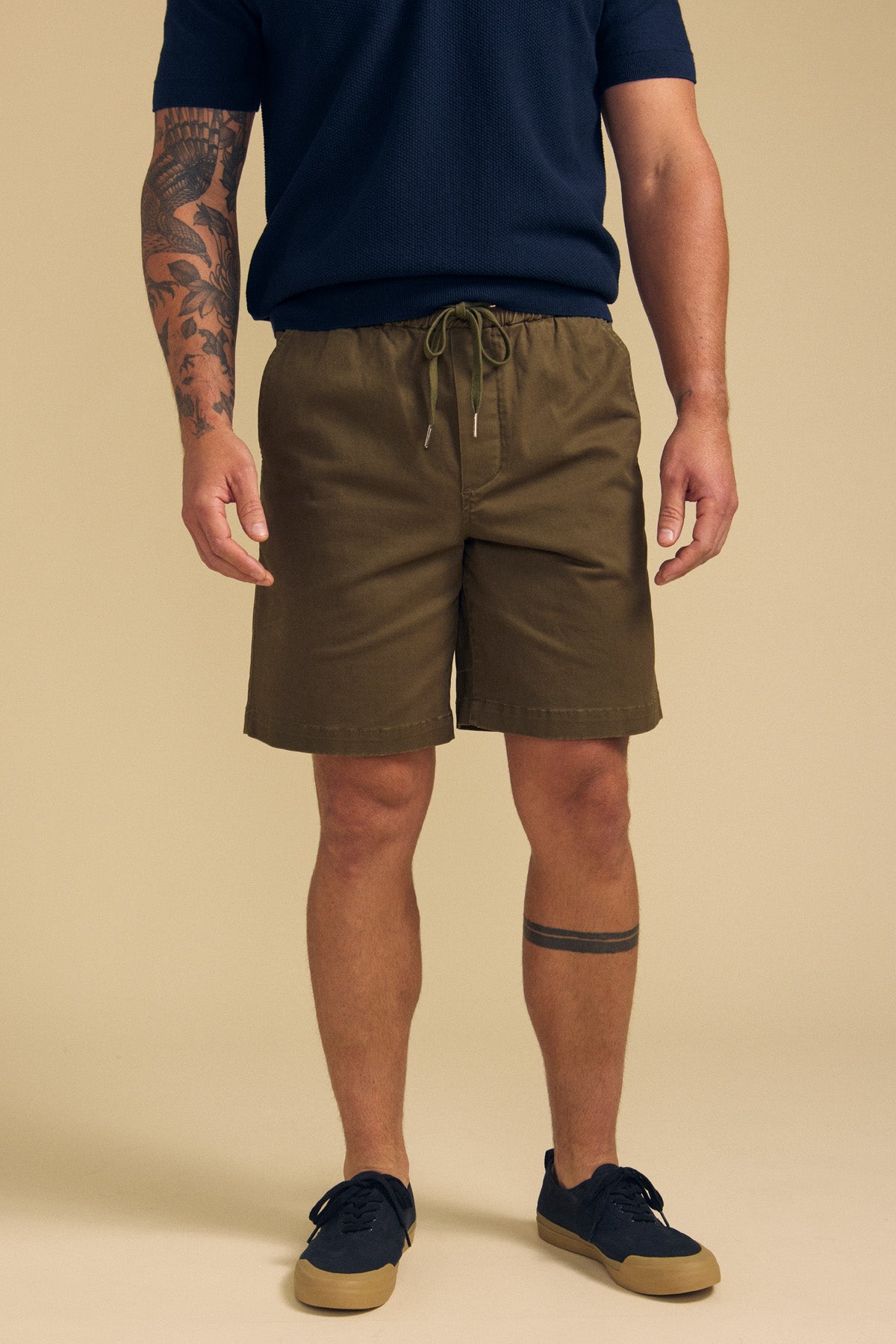 A man stands against a plain background wearing GARTH SHORTS from Velvet by Graham & Spencer, a navy short-sleeve shirt, and navy sneakers. Tattoos are visible on his left arm and right leg.