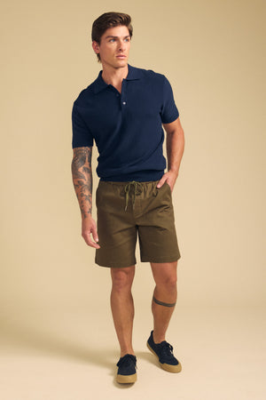 A man stands against a plain beige background wearing olive green Velvet by Graham & Spencer GARTH SHORTS, a navy polo shirt, and navy sneakers.
