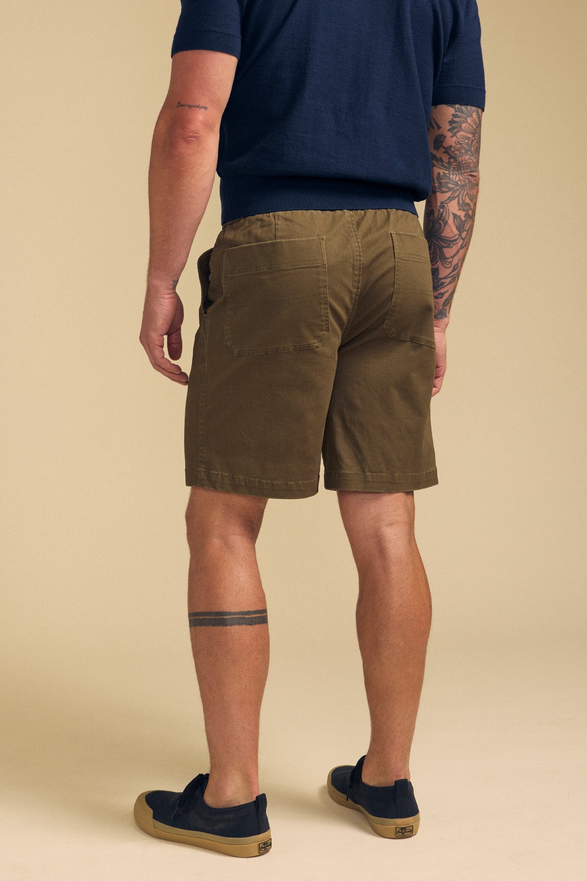 A man faces away, wearing Velvet by Graham & Spencer's GARTH SHORT in olive green, a navy shirt, black slip-ons, and showing tattoos on his left arm and right leg.