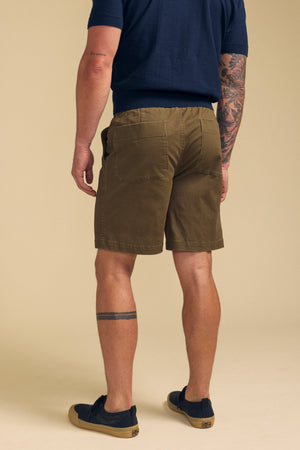 A man faces away, wearing Velvet by Graham & Spencer's GARTH SHORT in olive green, a navy shirt, black slip-ons, and showing tattoos on his left arm and right leg.