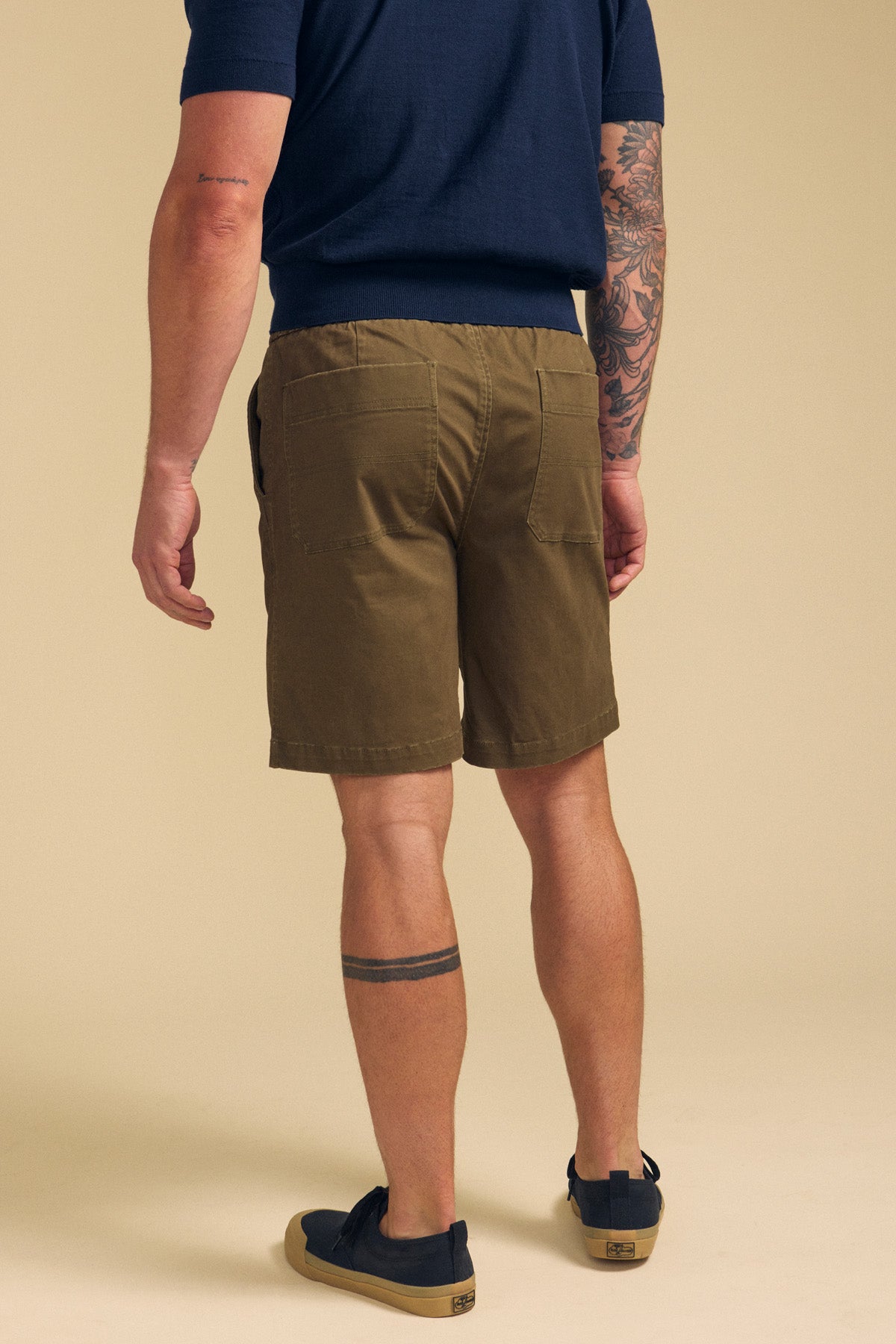 A man stands facing away against a plain beige background, wearing Velvet by Graham & Spencer’s GARTH SHORT in olive green, a navy shirt, and dark shoes. Tattoos are visible on his left arm and lower right leg.-41702009602241