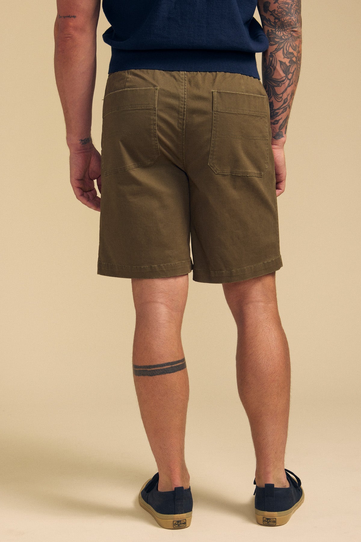 A person is shown from the back, standing on a beige background, wearing Velvet by Graham & Spencer GARTH SHORT in olive green, a navy shirt, and navy slip-on shoes. Visible tattoos are on their left arm and right leg.