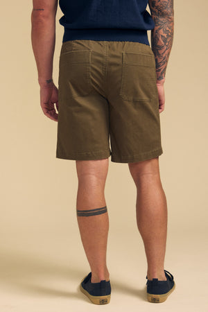 A person is shown from the back, standing on a beige background, wearing Velvet by Graham & Spencer GARTH SHORT in olive green, a navy shirt, and navy slip-on shoes. Visible tattoos are on their left arm and right leg.