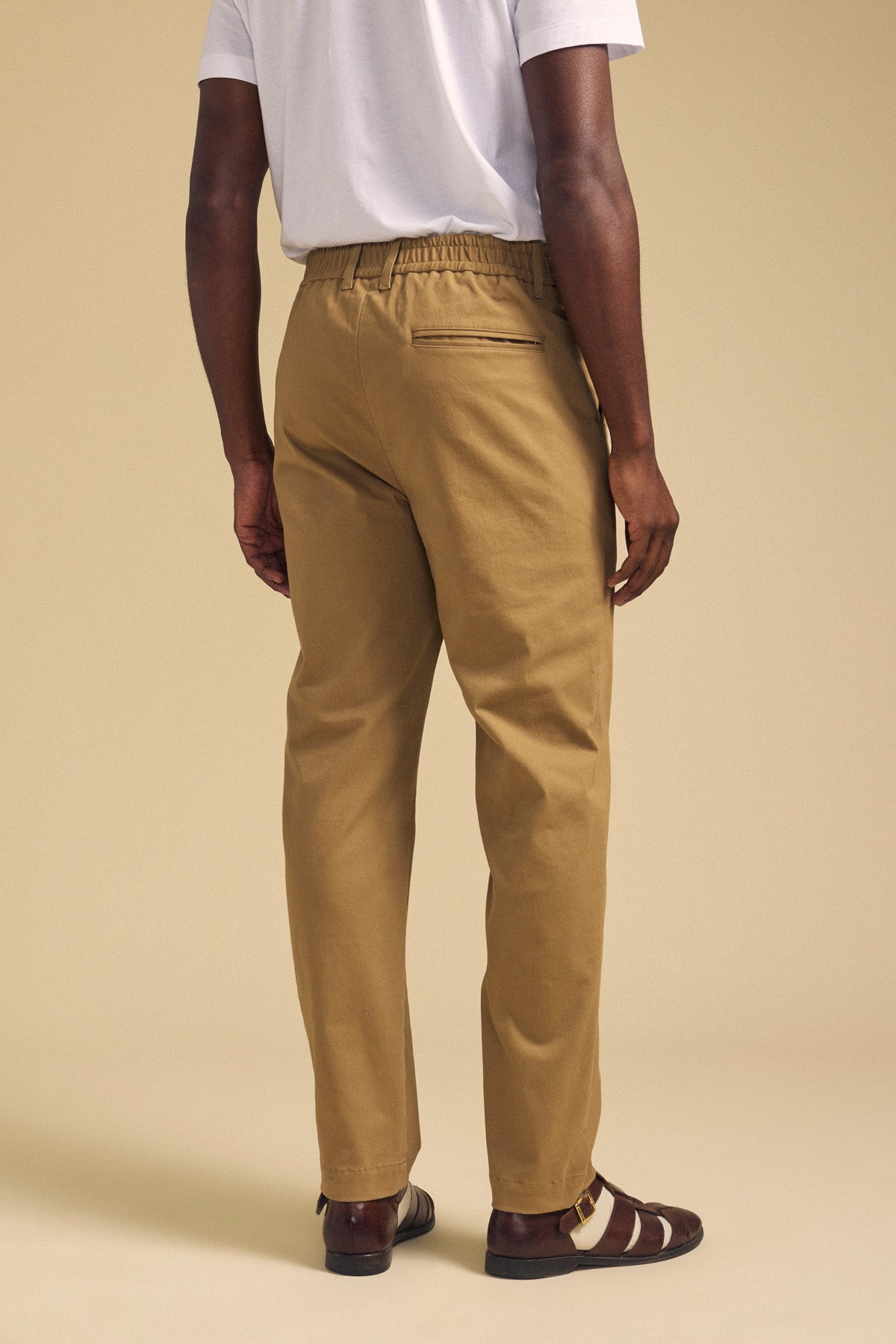 A person is seen from the back against a beige background, wearing a white t-shirt, brown sandals, and Velvet by Graham & Spencer's JUDE CHINO tan pants with an elastic waistband.