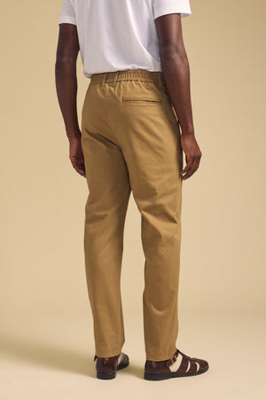A person is seen from the back against a beige background, wearing a white t-shirt, brown sandals, and Velvet by Graham & Spencer's JUDE CHINO tan pants with an elastic waistband.