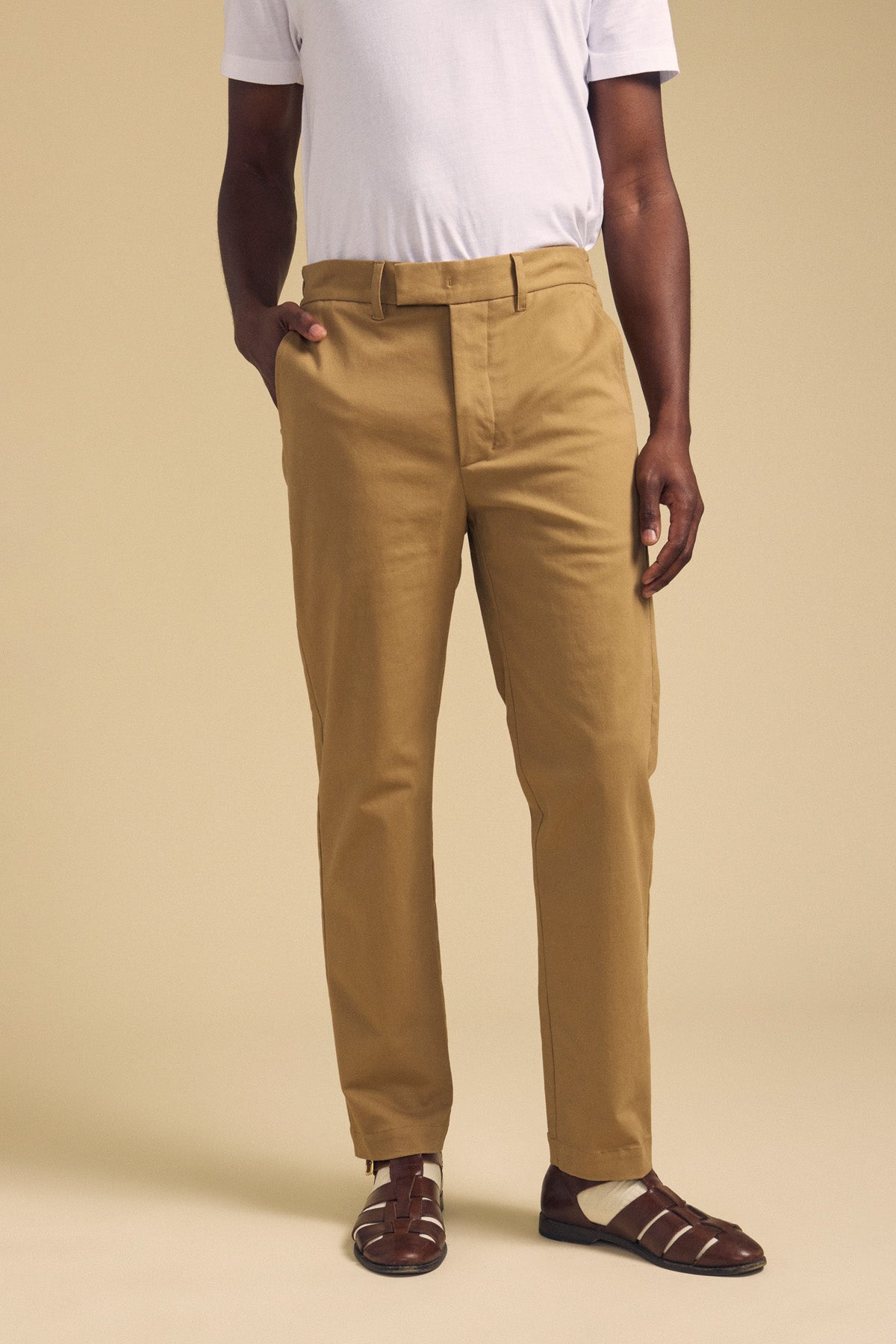 A person wears the JUDE CHINO by Velvet by Graham & Spencer, paired with a white t-shirt and brown sandals, standing against a beige background with one hand in their pocket.