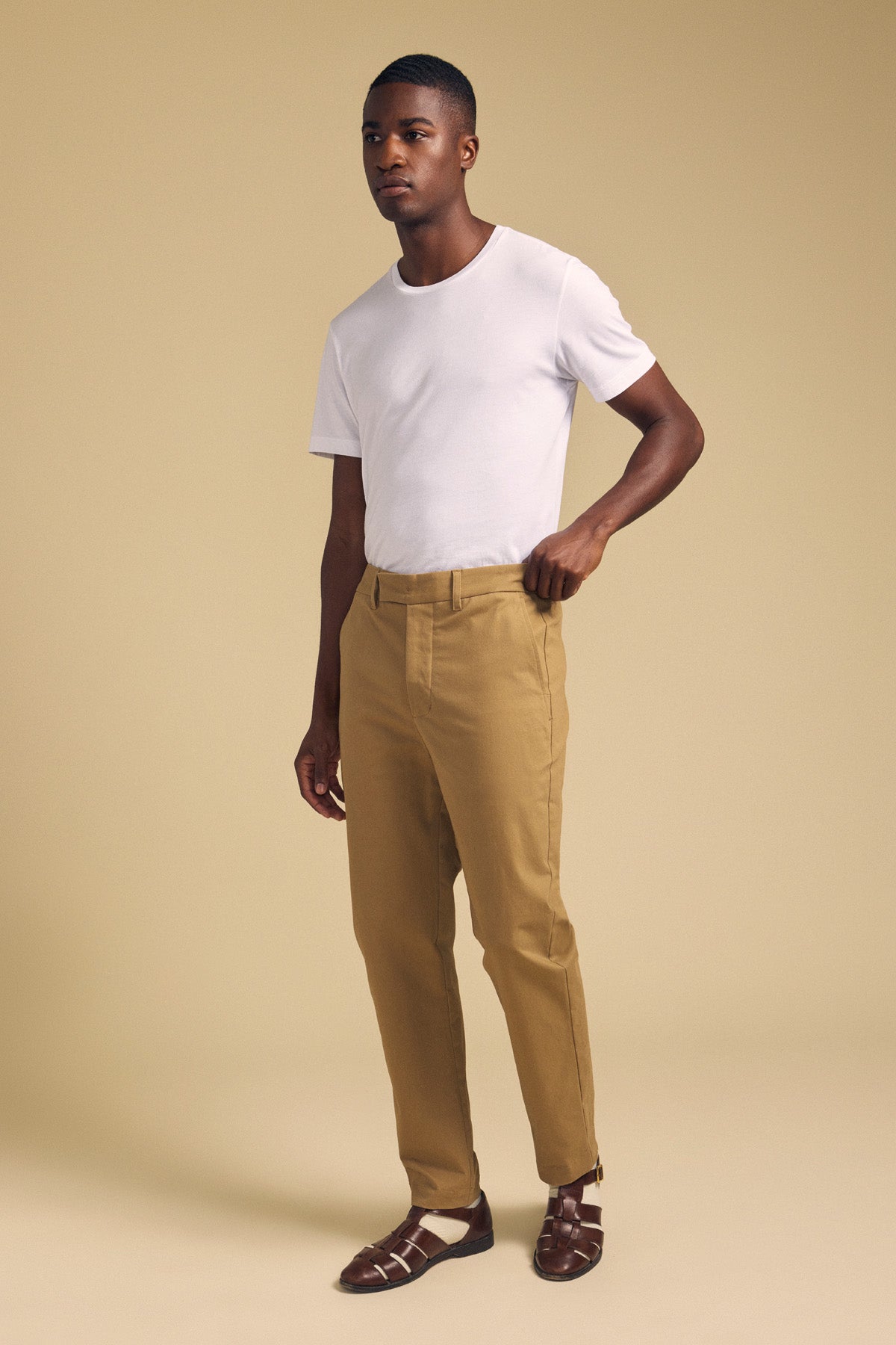 A man stands against a beige background wearing a white t-shirt, brown sandals, and JUDE CHINO pants by Velvet by Graham & Spencer.