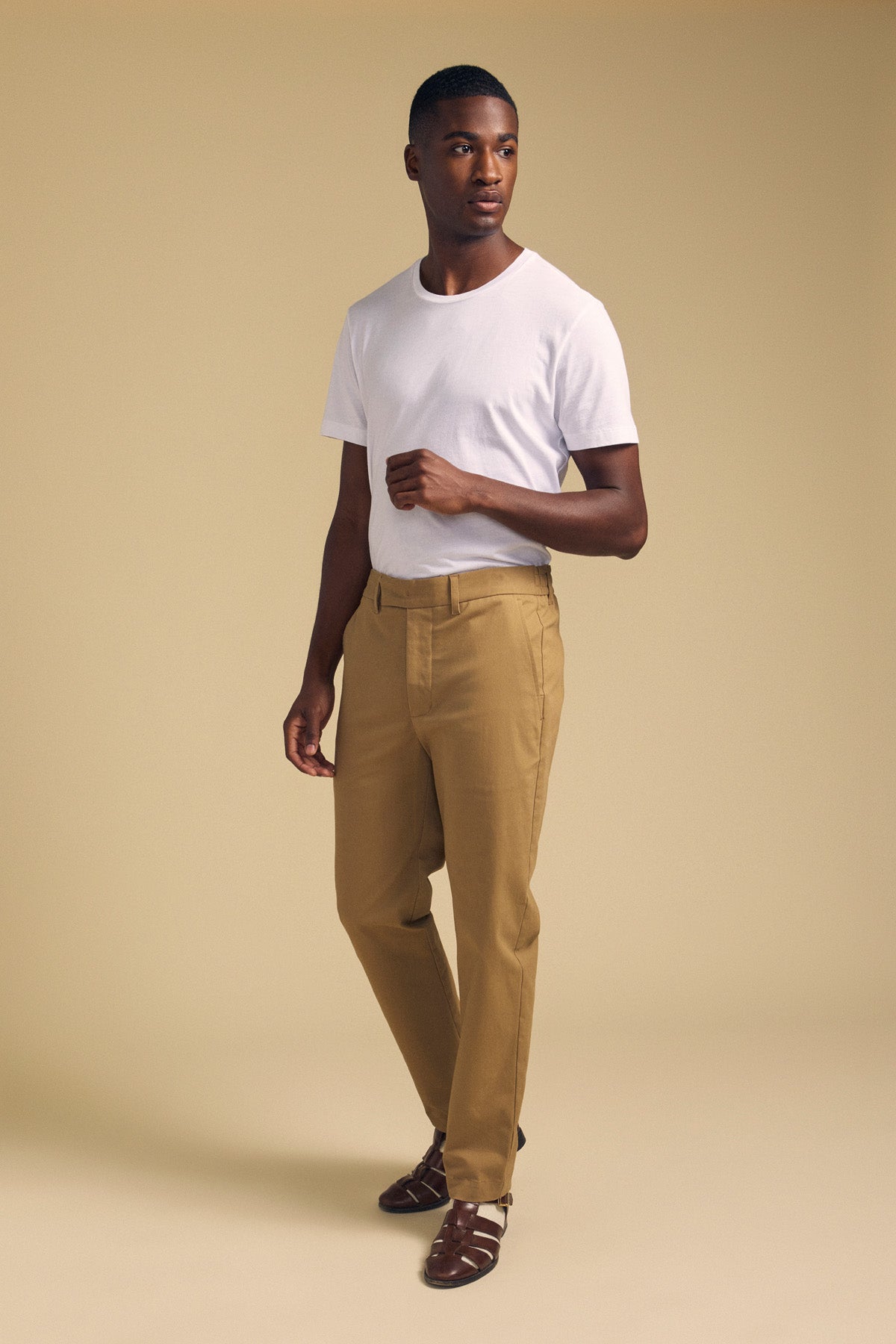 A man stands against a beige background wearing a white t-shirt, brown sandals, and JUDE CHINO trousers by Velvet by Graham & Spencer.