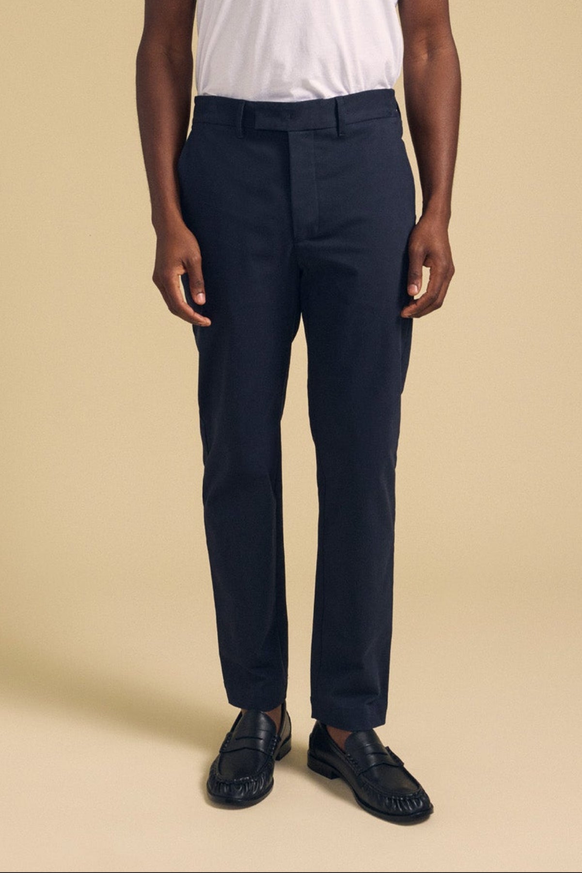 A person wearing Velvet by Graham & Spencer’s JUDE CHINO stands against a plain beige background, paired with a white t-shirt and black loafers.-41702028017857