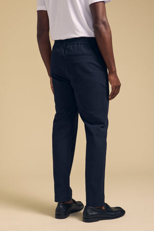 A person is seen from the back against a beige background, wearing Velvet by Graham & Spencer's JUDE CHINO in navy blue with an elastic waistband, paired with a white t-shirt and black loafers.