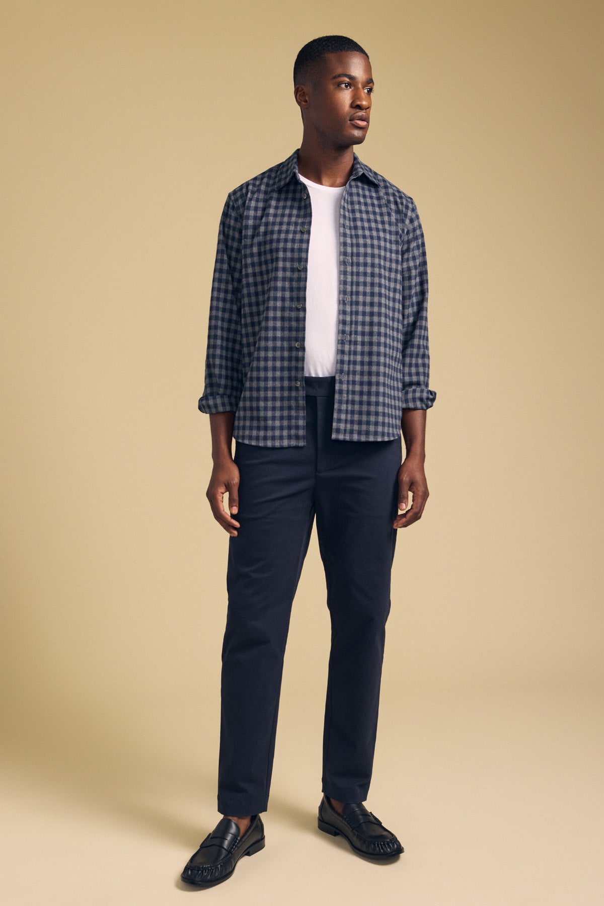 A man stands against a beige background wearing Velvet by Graham & Spencer's JUDE CHINO, paired with a blue checked shirt over a white t-shirt and black shoes.-41702027591873