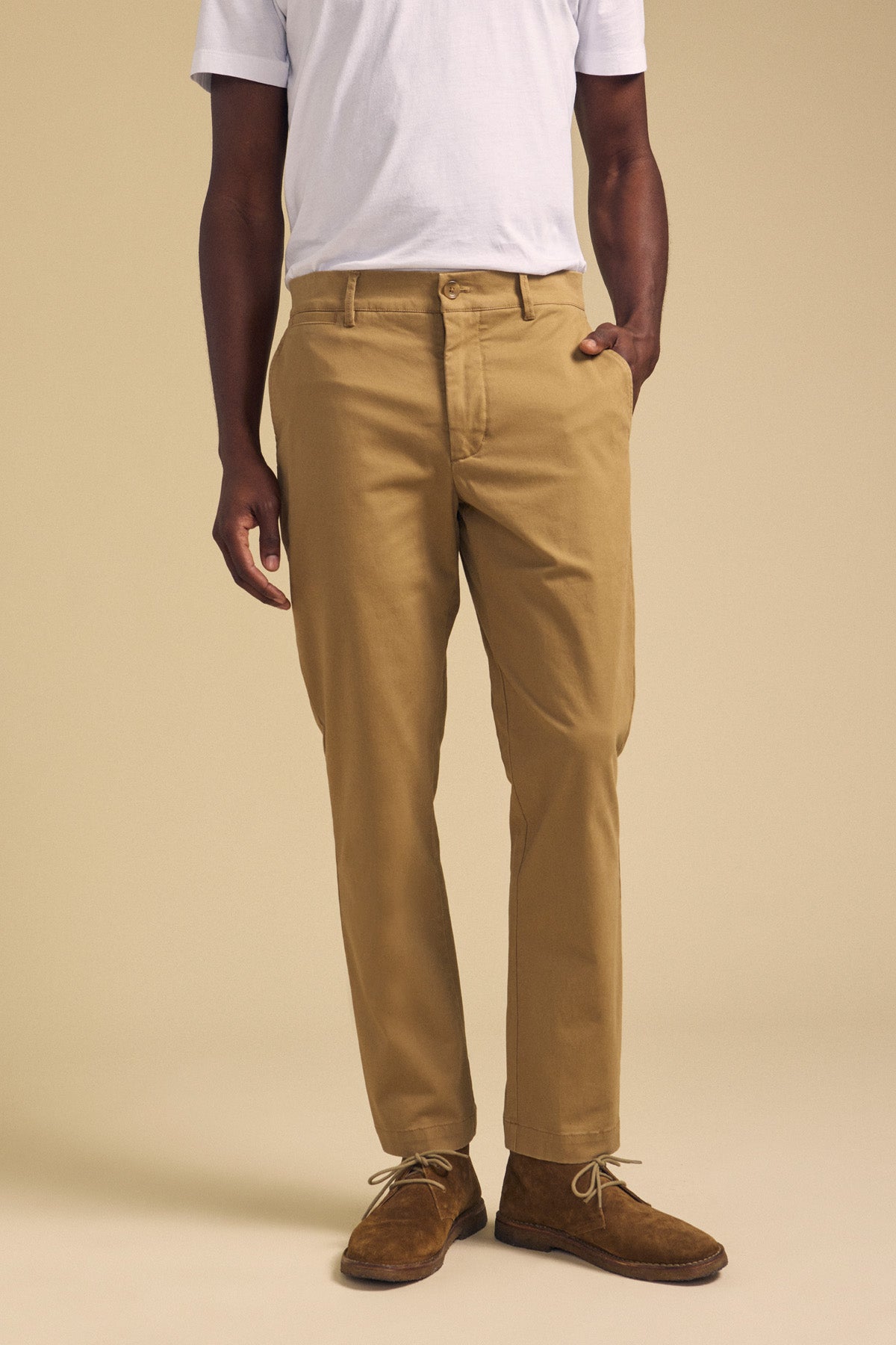 A person wearing a white t-shirt, the LUIS CHINO by Velvet by Graham & Spencer, and brown suede shoes stands against a beige background with one hand in their pocket.