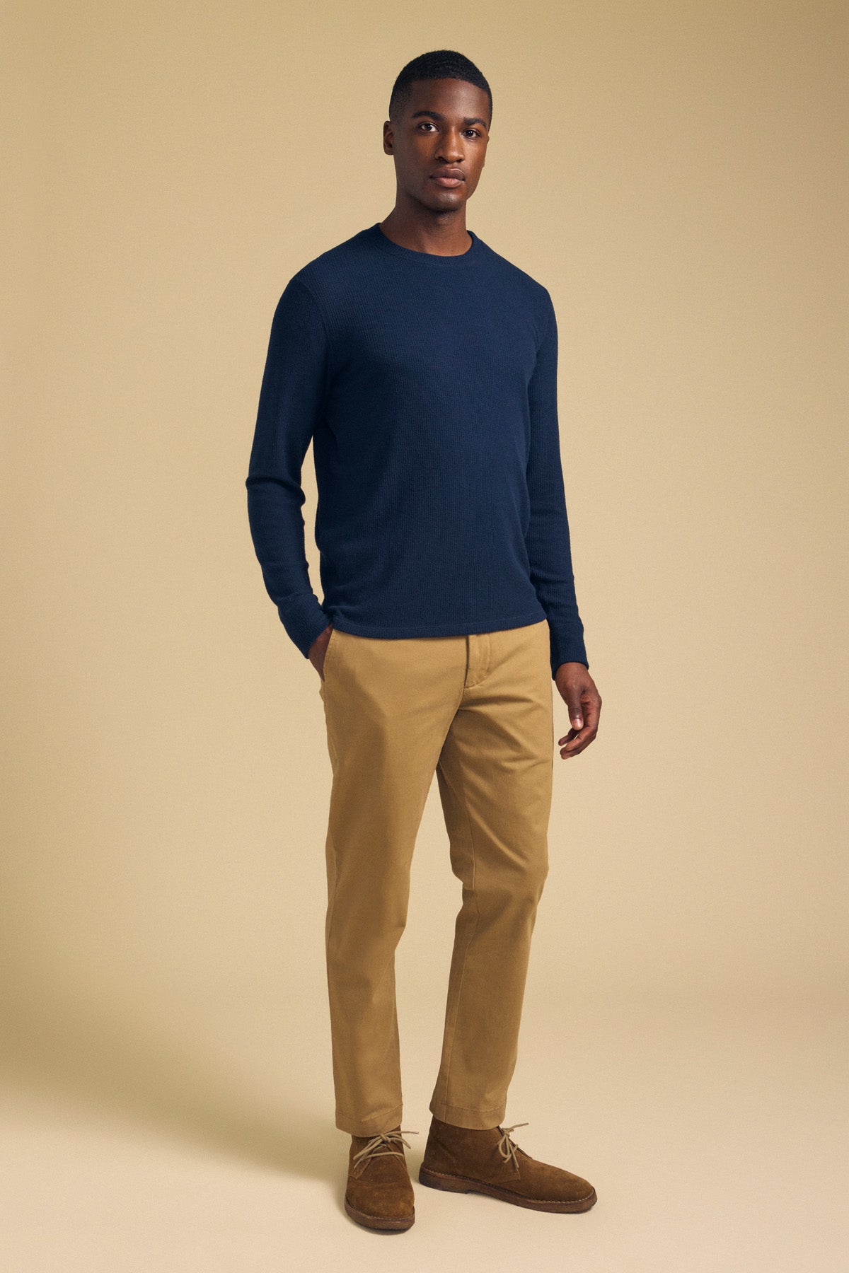 A man stands against a beige background wearing the LUIS CHINO pants by Velvet by Graham & Spencer, paired with a navy long-sleeve shirt and brown shoes, with his left hand in his pocket.