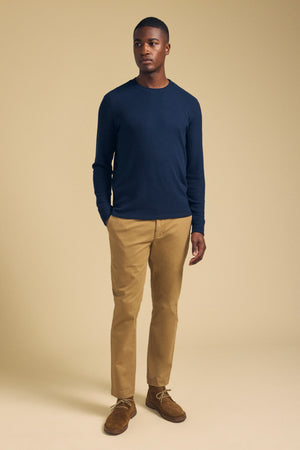 A man stands against a beige background wearing Velvet by Graham & Spencer’s LUIS CHINO pants in tan, paired with a navy blue long sleeve shirt and brown shoes, looking slightly to the side with his hands in his pockets.