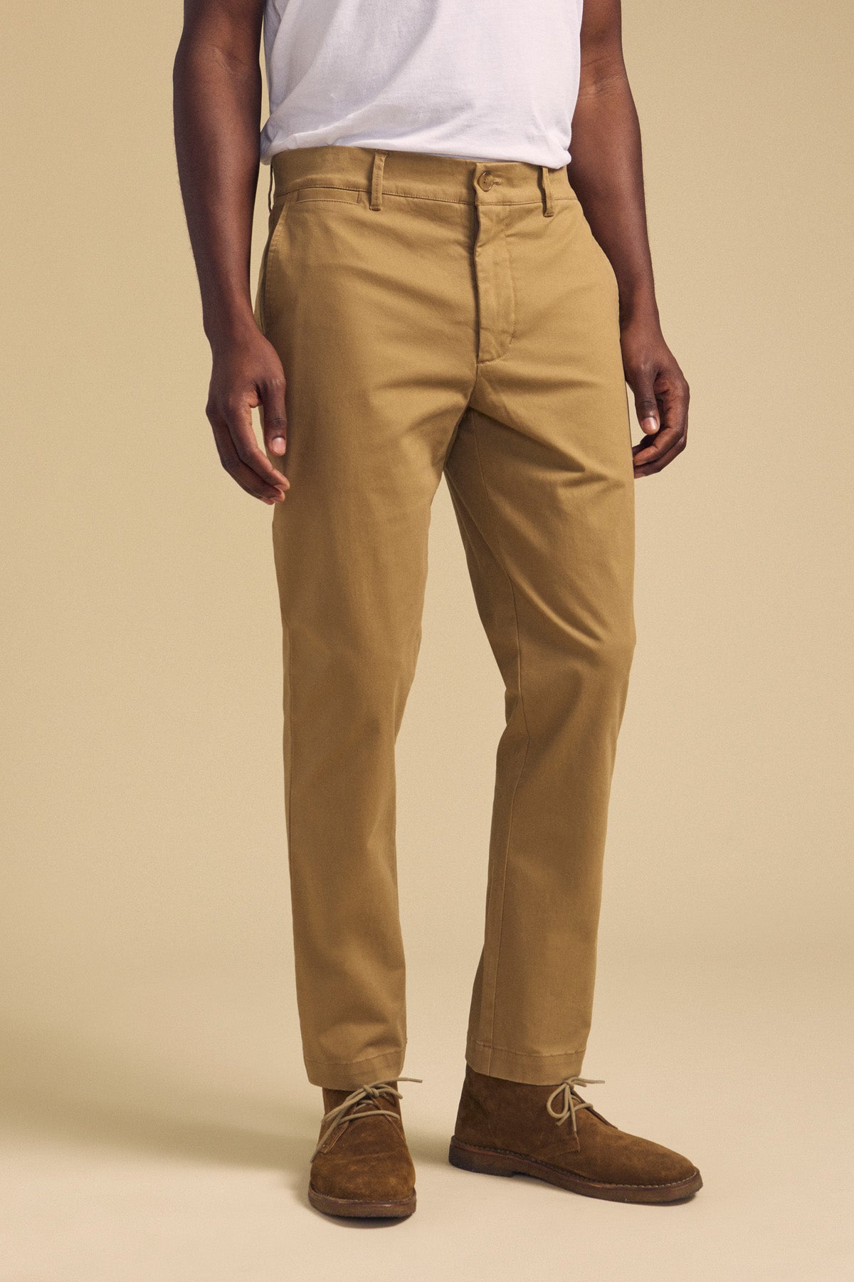 A person wearing the LUIS CHINO pants by Velvet by Graham & Spencer, paired with a white shirt and brown suede shoes, stands against a plain beige background.