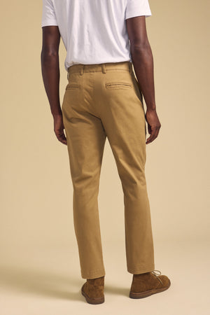 A person stands facing away against a beige background, wearing Velvet by Graham & Spencer’s LUIS CHINO tan pants with belt loops and back pockets, paired with a white T-shirt and brown suede shoes.