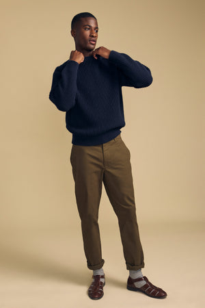 A man stands against a beige background wearing the LUIS CHINO khaki pants with cuffs by Velvet by Graham & Spencer, a navy sweater, gray socks, and brown dress shoes, holding the collar of his sweater with both hands.