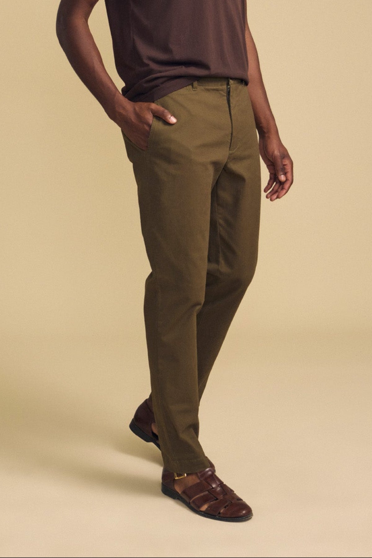 Clad in the LUIS CHINO by Velvet by Graham & Spencer, this individual pairs olive green trousers with a dark brown shirt and brown sandals, standing with one hand in their pocket against a beige background.